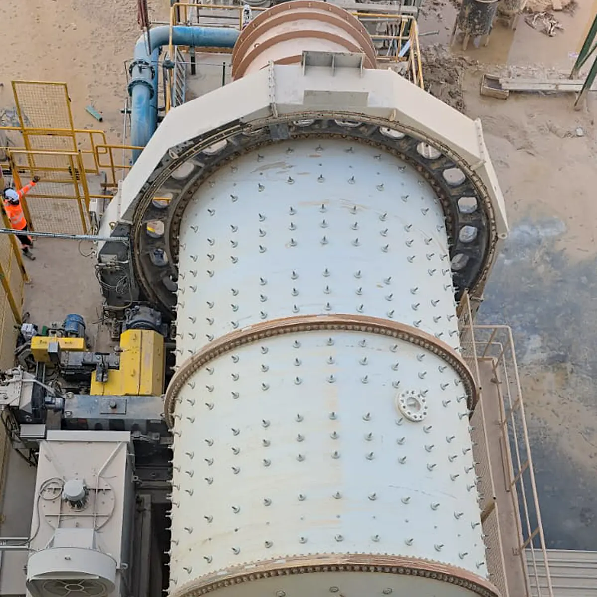 Large industrial ball mill grinding machine with metal bolts and adjacent blue pipe in a dusty industrial site.