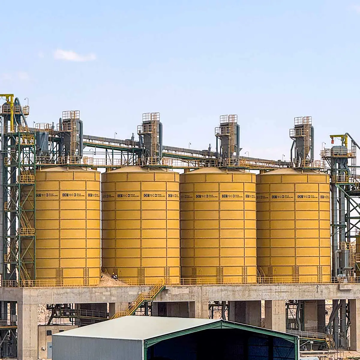Four large yellow cylindrical industrial storage silos with metal walkways and staircases against a clear sky.