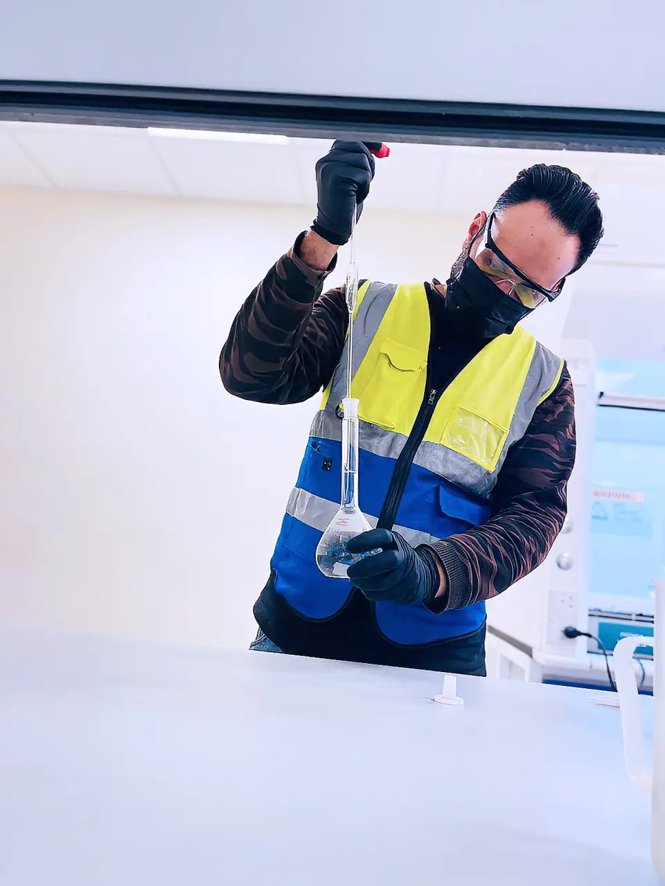 Lab technician wearing safety goggles, gloves, and a high-visibility vest carefully pouring liquid into a volumetric flask in a laboratory.