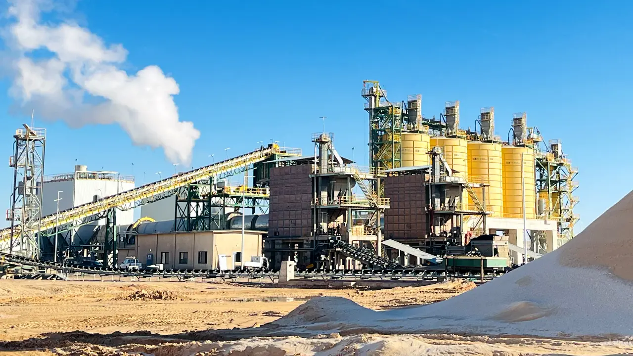 Dry phosphate concentrate storage area with conveyor belts, large storage silos, and smoke emission under a clear blue sky.