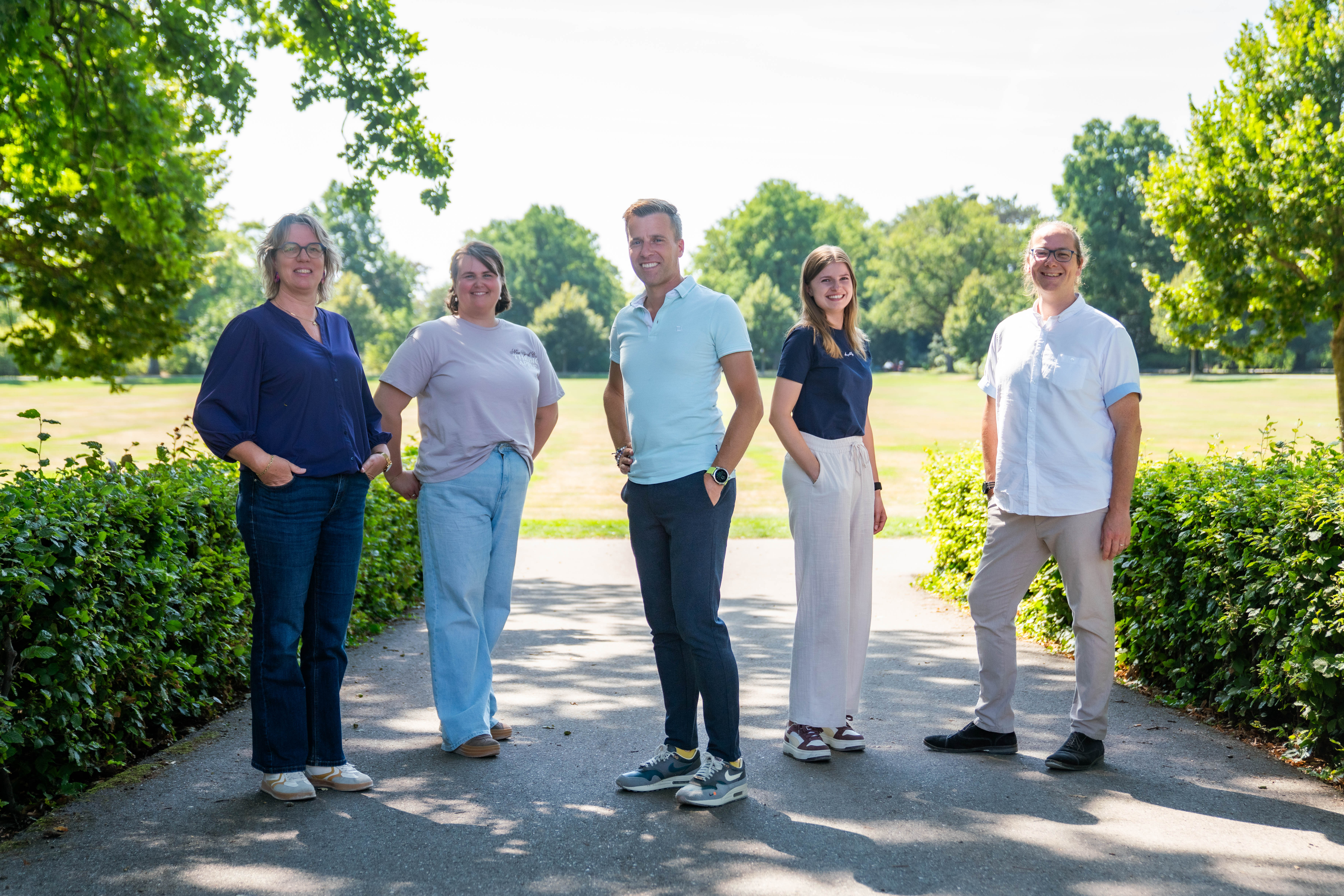 Group of five adults standing on a paved path in a park with green bushes and trees, smiling and posing for the camera on a sunny day.