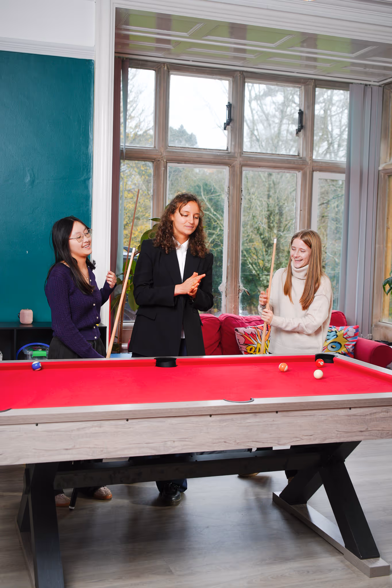 Three young women playing pool on a red-felt billiards table in a room with large windows and colorful pillows.