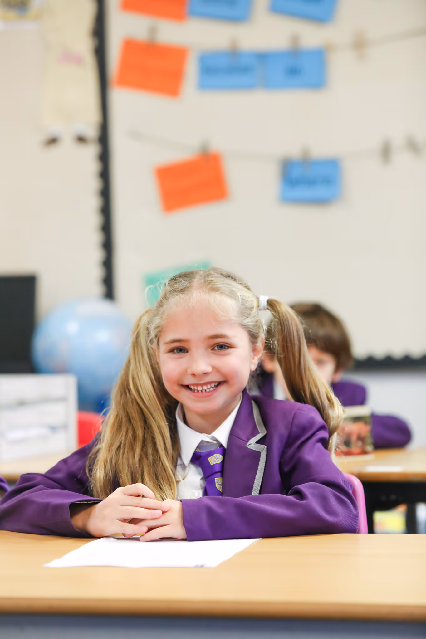 Smiling schoolgirl with blonde pigtails wearing a purple blazer and tie, seated at a classroom desk.