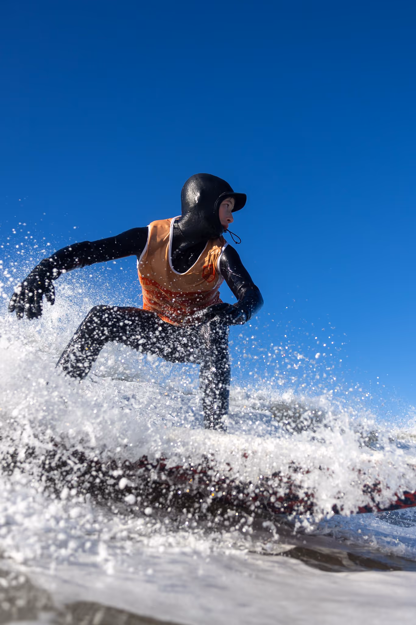 Surfer in a black wetsuit and orange vest riding a wave with water splashing around under a clear blue sky.