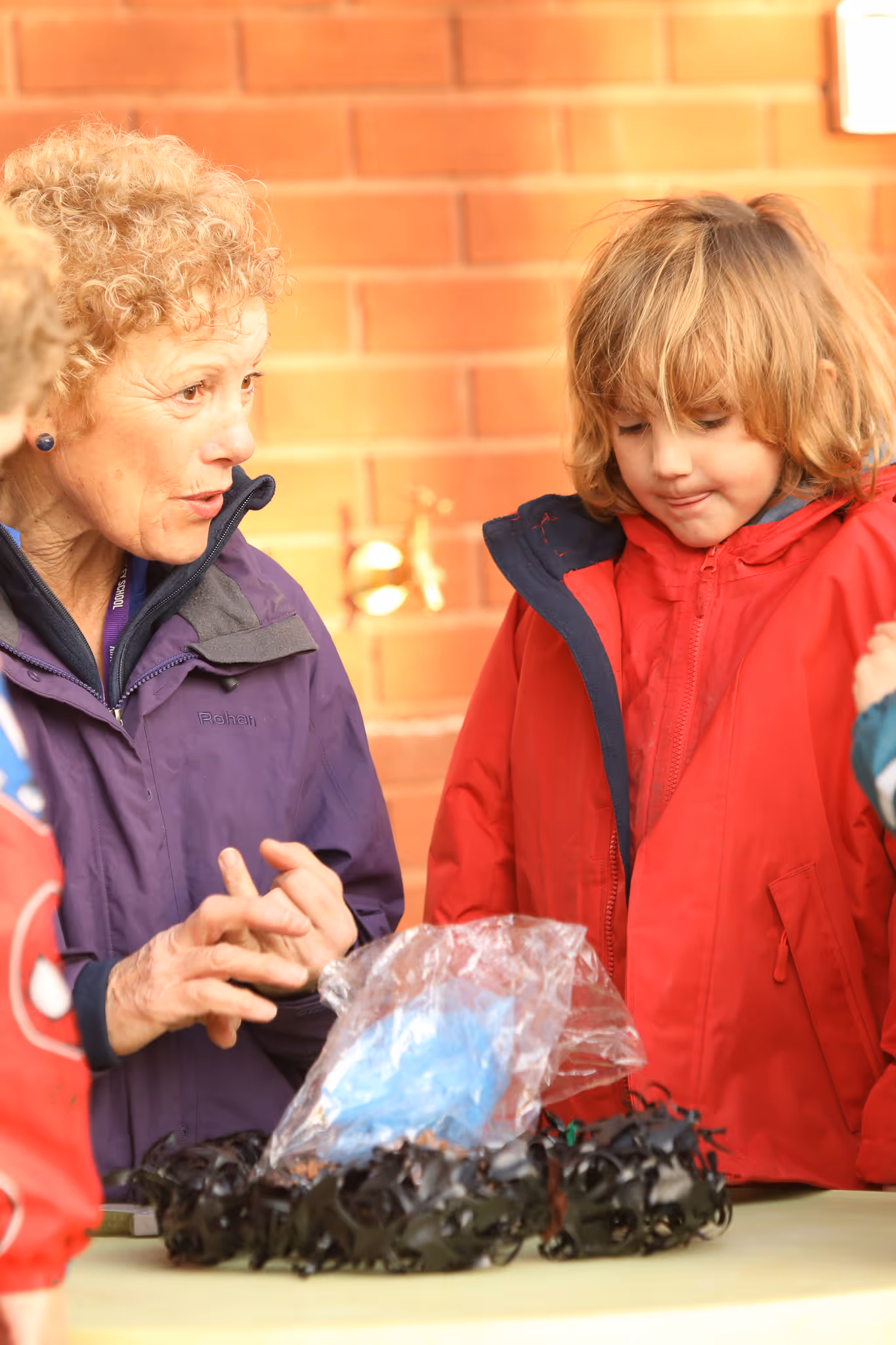 Older woman explaining something to a child in a red jacket at a table with a plastic-wrapped object and black material.