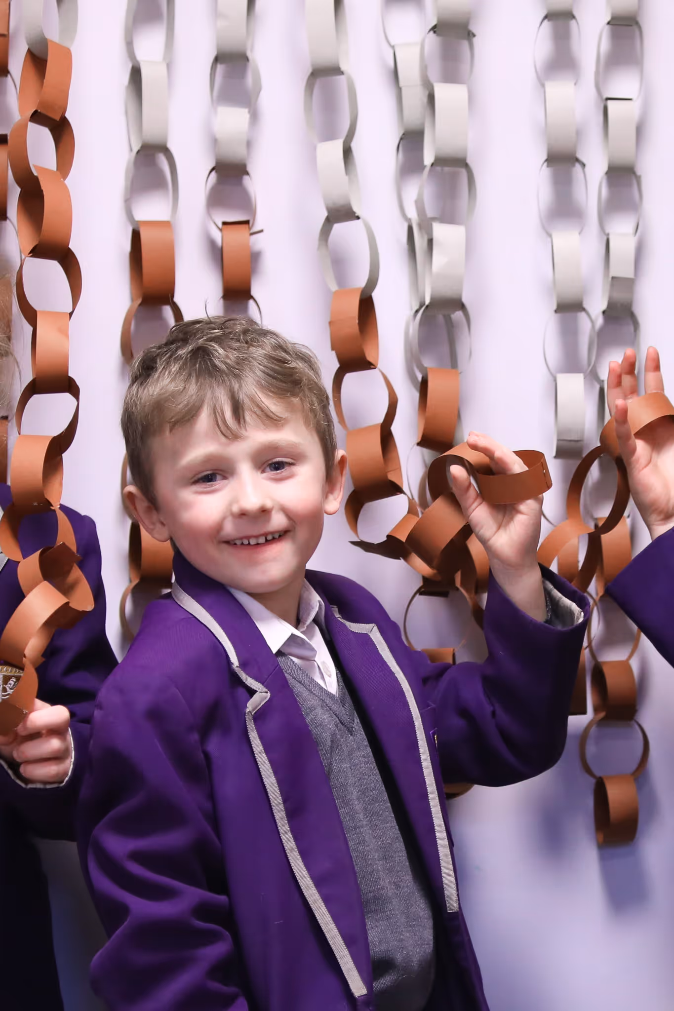 Smiling young boy in a purple school blazer holding a brown paper chain decoration.