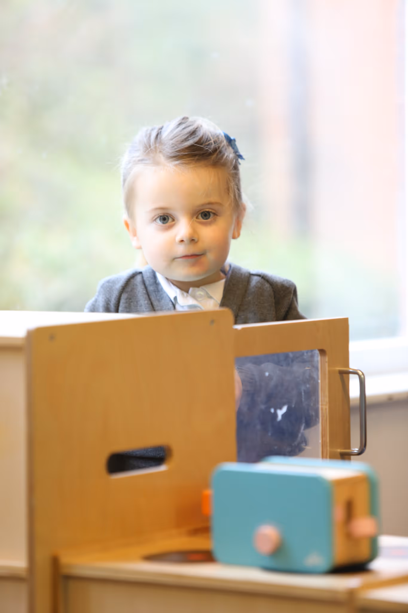 Young child with light hair and a blue bow playing with a wooden toy kitchen near a window.