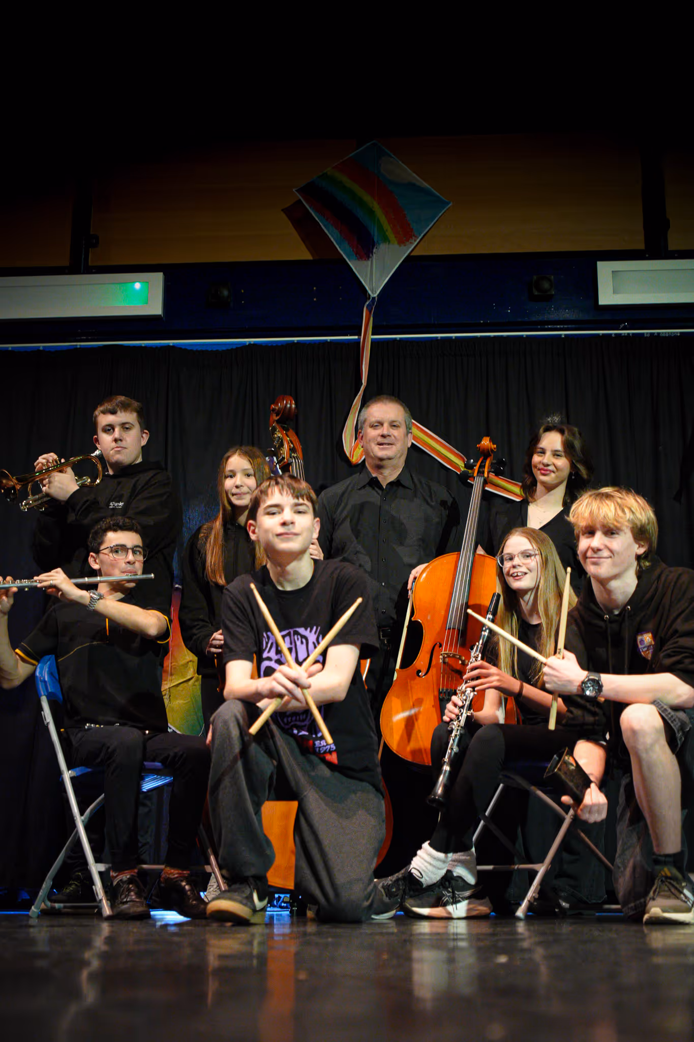 Group of seven young musicians and one adult posing with instruments including flute, trumpet, cello, clarinet, and drumsticks on stage with a rainbow kite decoration.