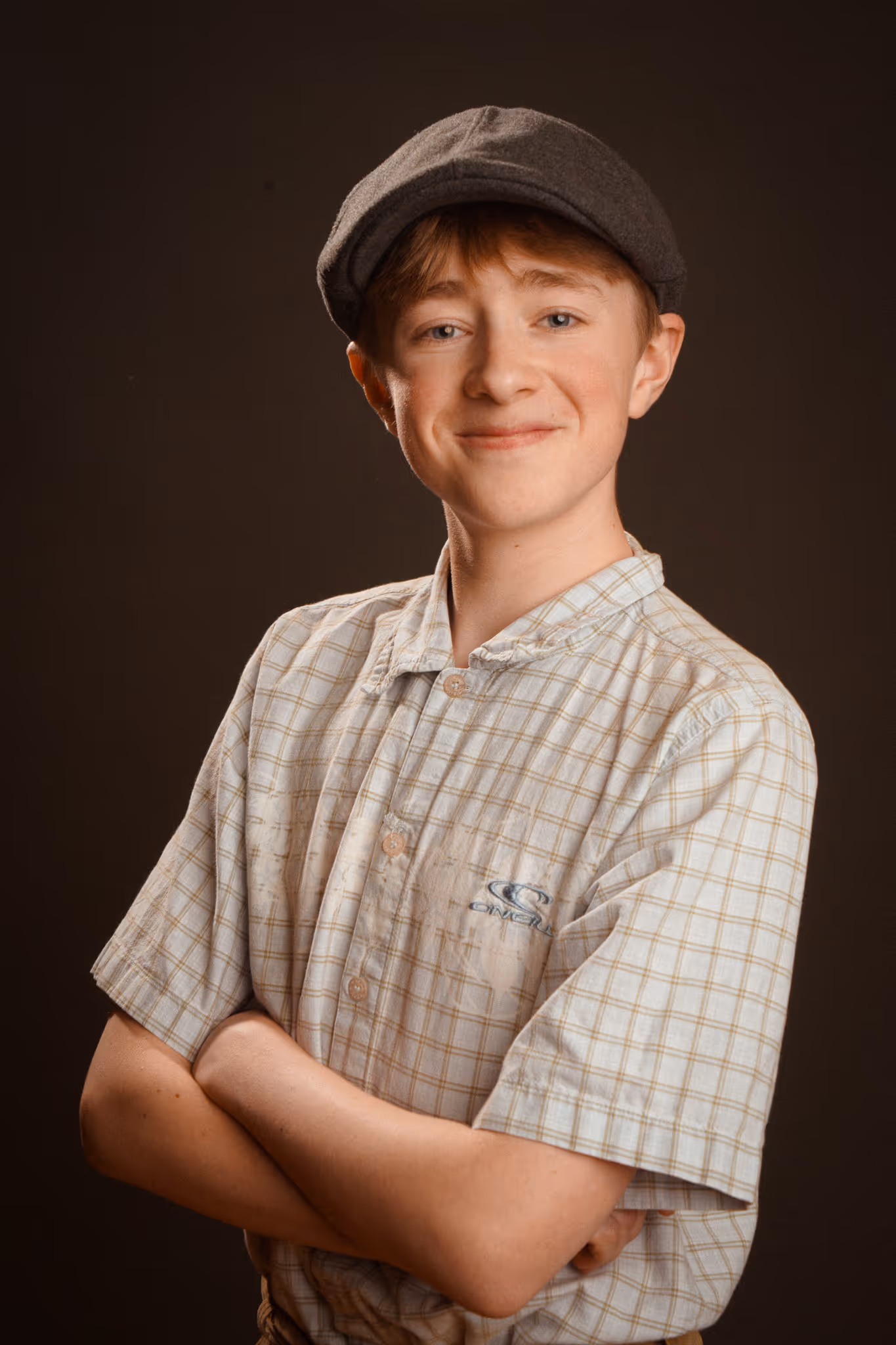 Smiling young boy with crossed arms wearing a gray flat cap and a checkered short-sleeve shirt against a dark background.