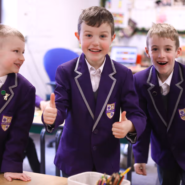 Three boys in purple school blazers smiling and giving thumbs up in a classroom.
