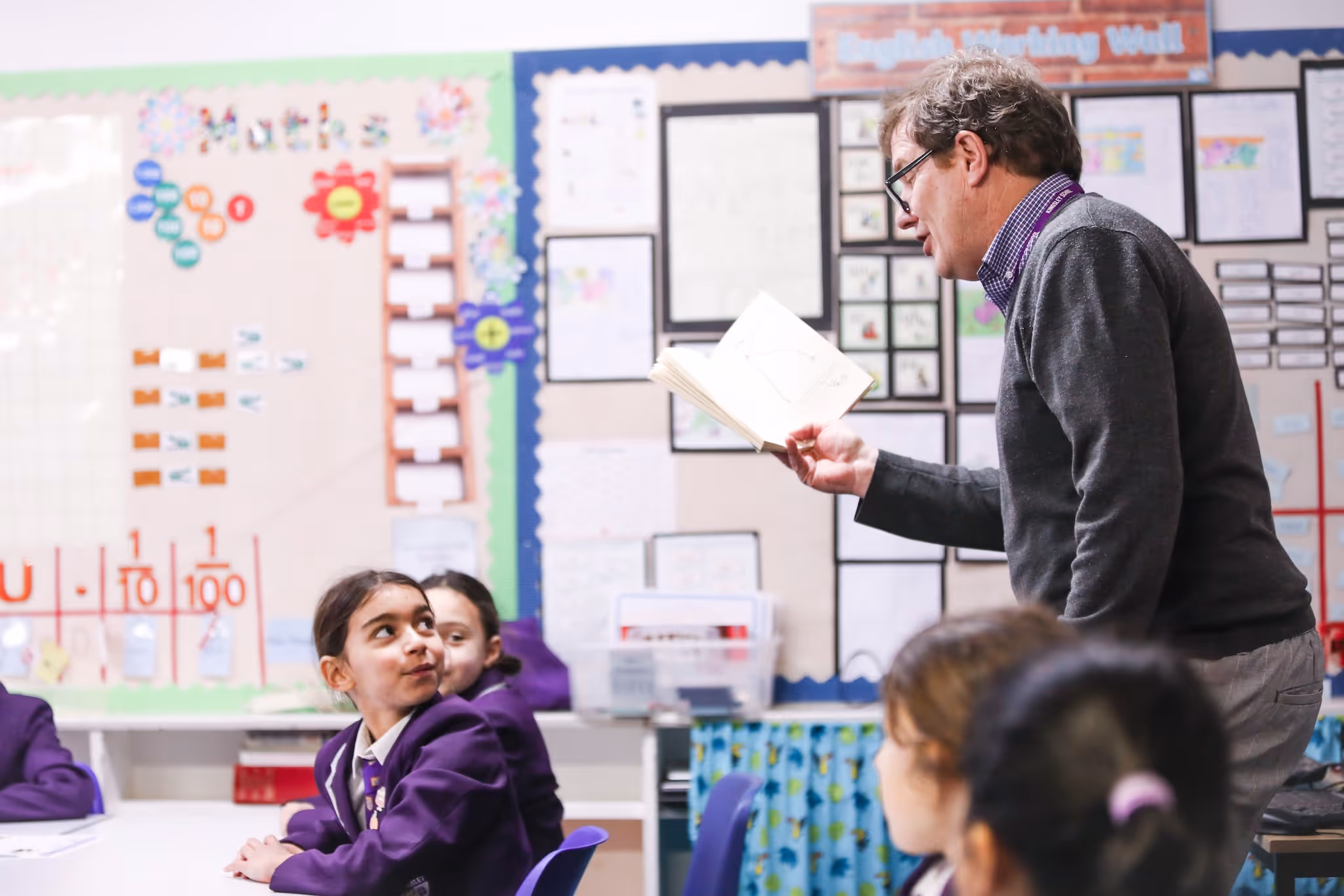 Male teacher reading from a book to attentive students in a classroom with colorful educational posters.