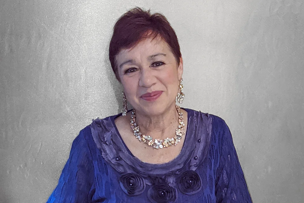 Found of TWL Logistics Wilhelmina Ligthart smiling in a purple dress and floral necklace in front of a gray background.