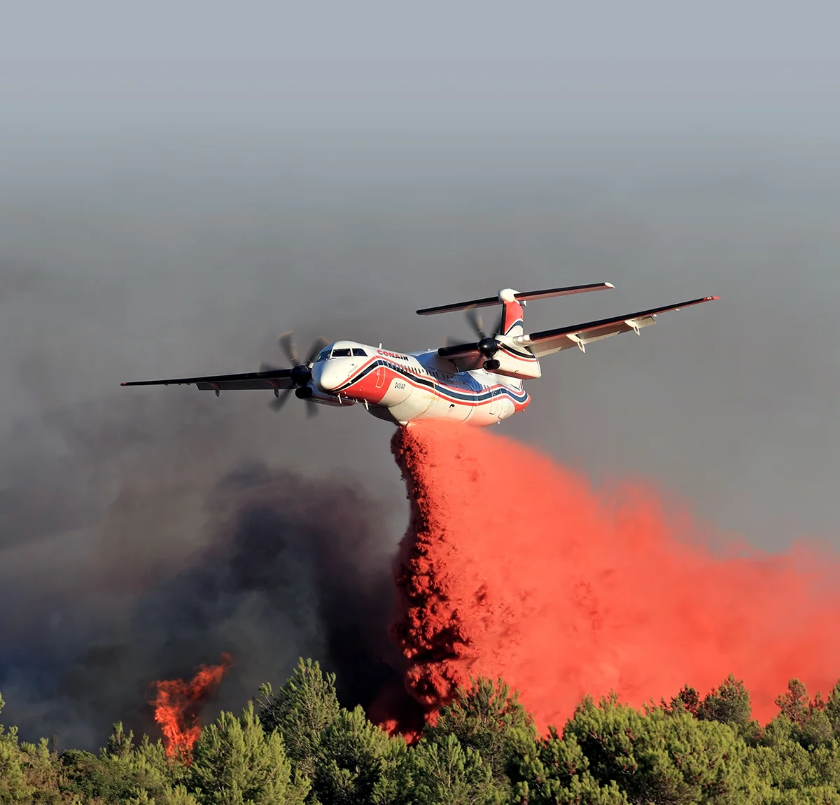 Conair Q400-AT firefighting aircraft drops red fire retardant over a forest fire amid smoke and flames.