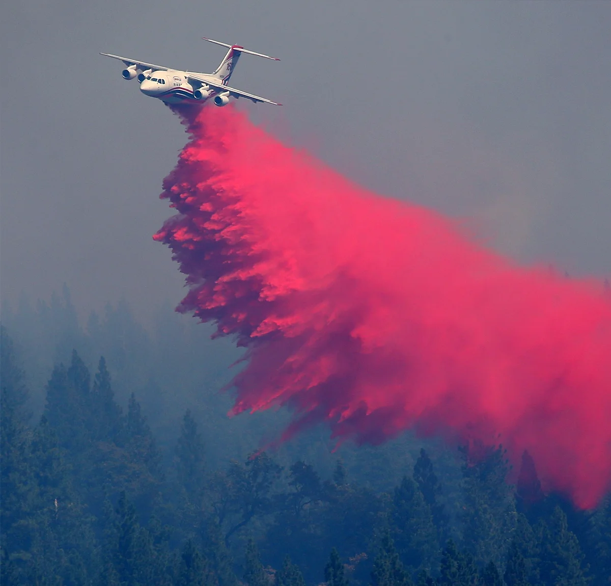 Avro RJ85 aircraft releasing red fire retardant over forest to combat wildfire smoke in the background.