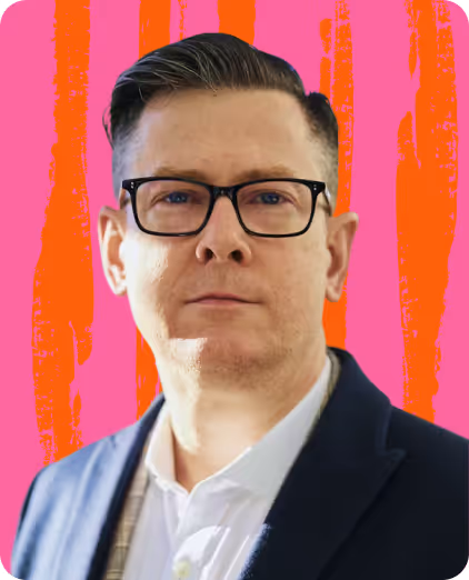 Man with short dark hair and glasses wearing a dark blazer and white shirt against a pink and orange striped background.