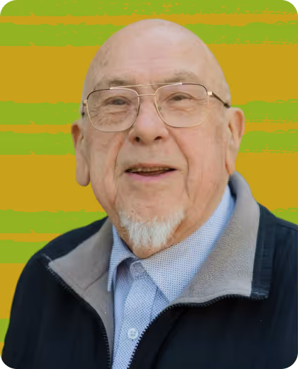 Elderly man with glasses and a white beard, smiling, wearing a light blue shirt and dark jacket.