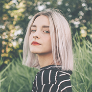 Young woman with blonde bob haircut wearing a black and white striped top standing outdoors with green foliage background.