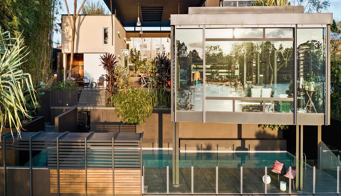 Modern multi-level house with large glass windows, greenery, outdoor seating, and a glass-fenced swimming pool.