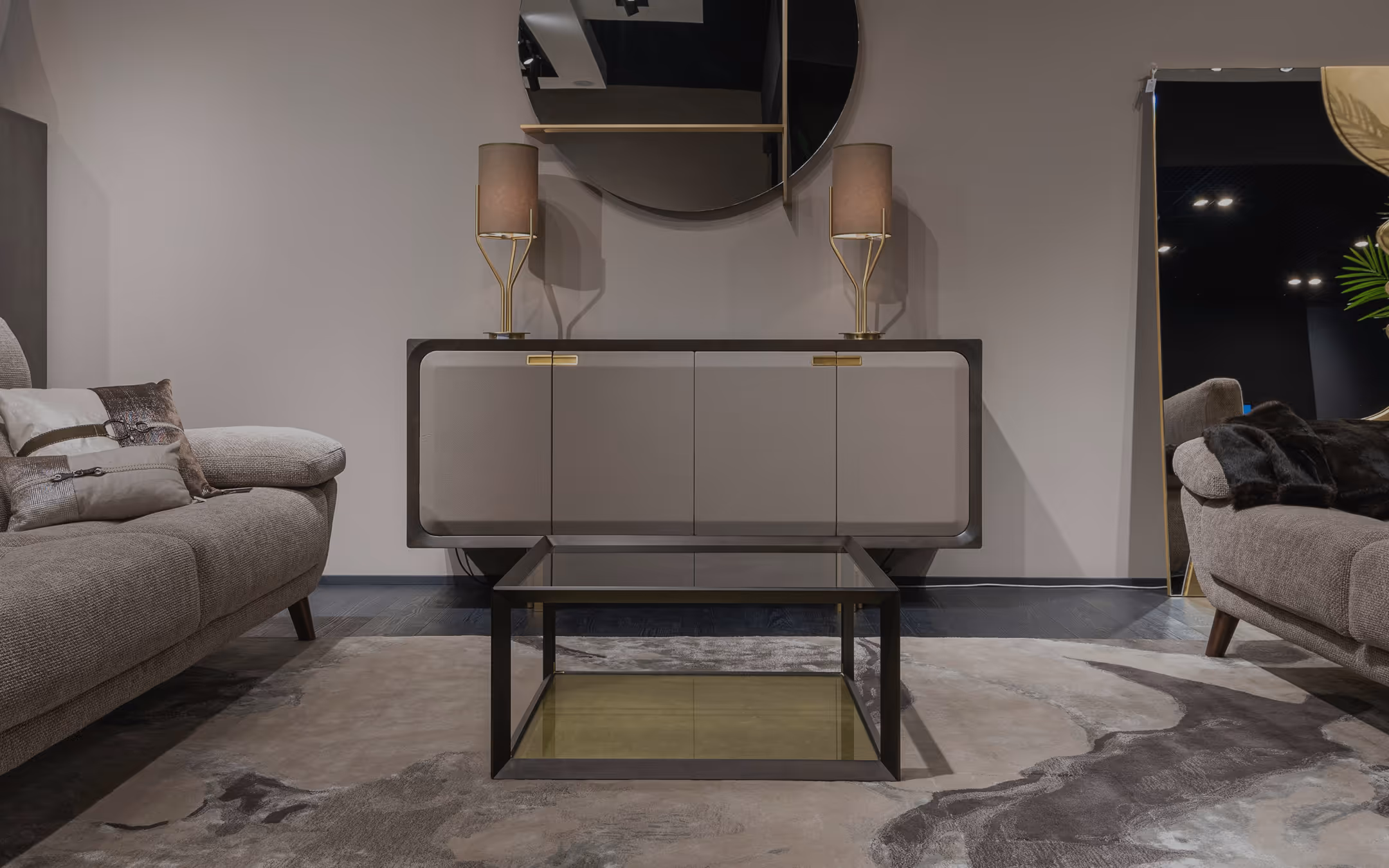 Modern living room with two gray fabric sofas, a square glass coffee table, a sideboard with gold-accented lamps, and a large round mirror on the wall.