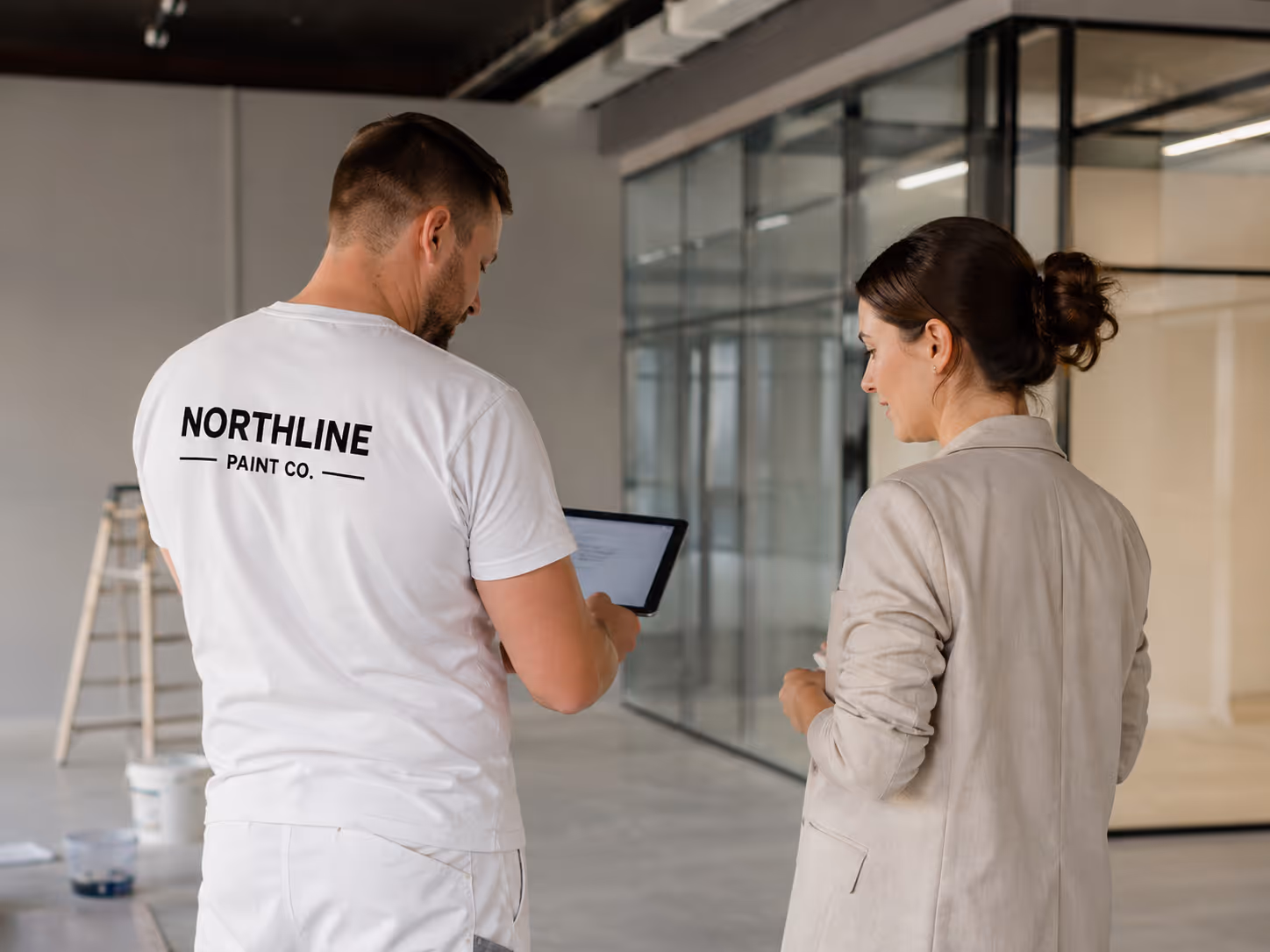 A man in a white t-shirt with 'Northline Paint Co.' on the back shows a tablet to a woman in a beige blazer inside a modern office space.