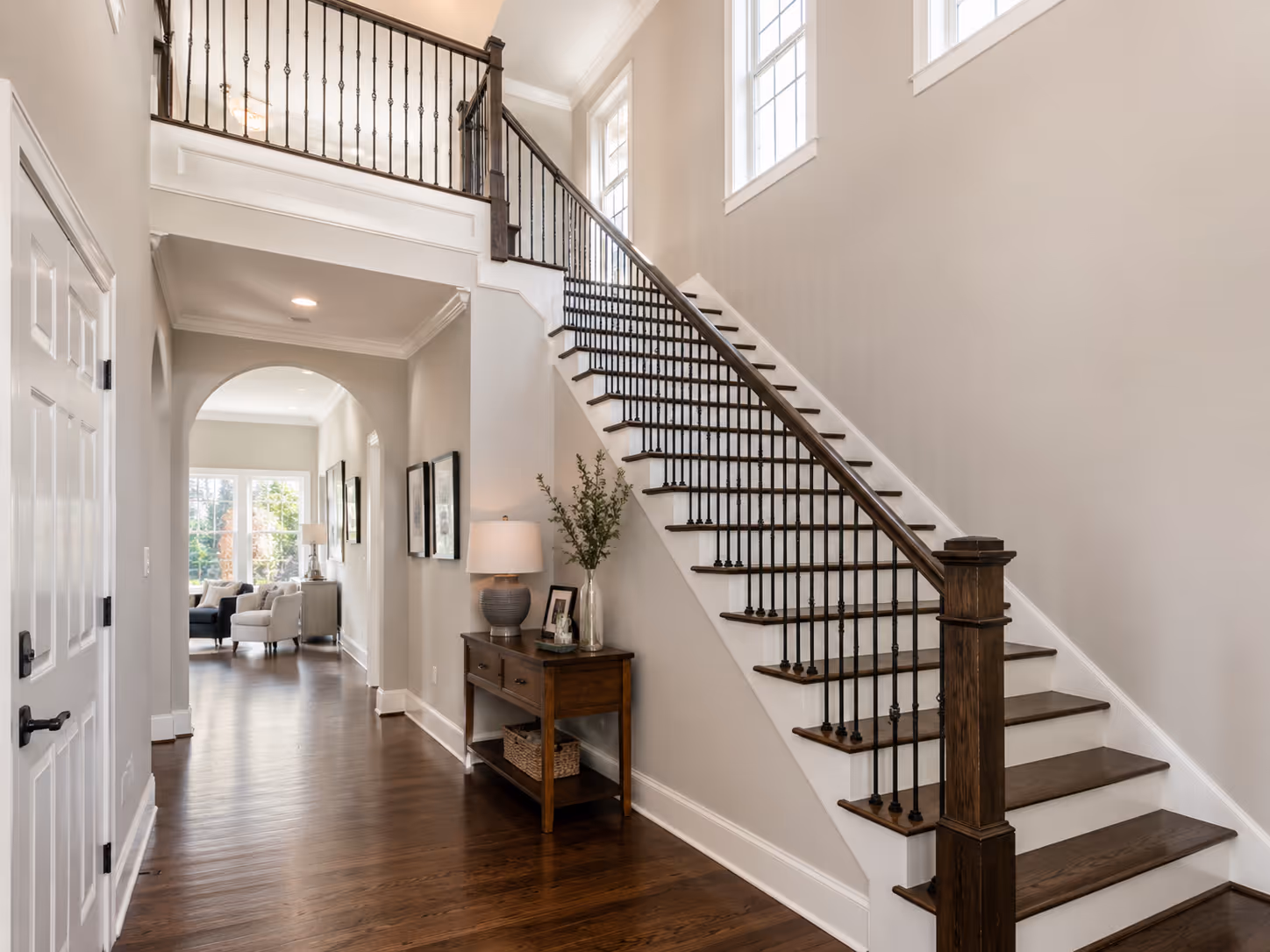 Bright hallway with wooden floor, white walls, dark wood staircase with metal balusters, and a console table with a lamp and plant.