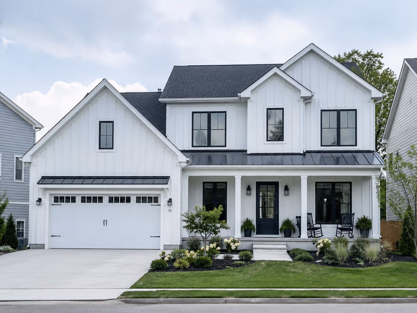 Modern two-story white house with black-trim windows, front porch with rocking chairs, attached garage, and neatly landscaped yard.