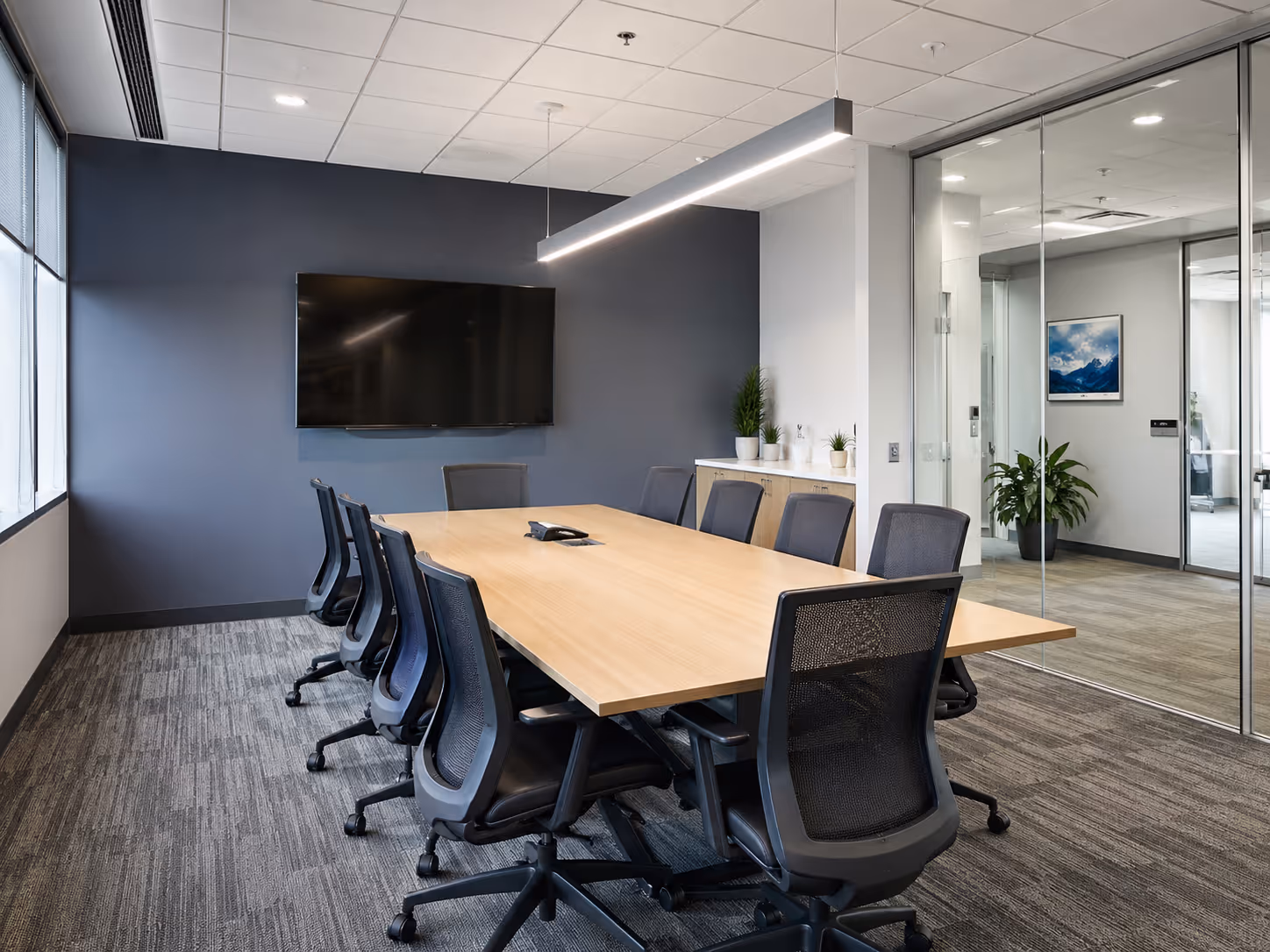 Modern conference room with a long wooden table surrounded by black mesh office chairs, a large wall-mounted TV, and glass walls.