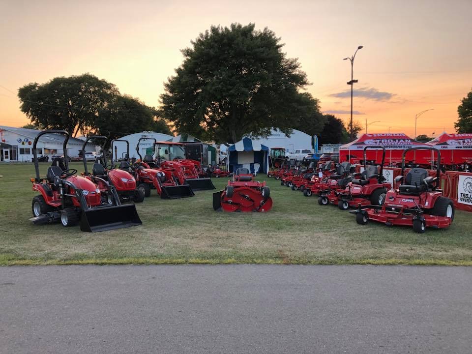 Outdoor display of red lawn tractors and zero-turn mowers arranged in rows on grass, with tents and large trees in the background at sunset.
