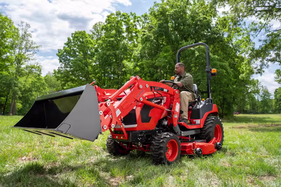 A man operating a red compact tractor equipped with a front loader attachment in a grassy field surrounded by trees under a partly cloudy sky.