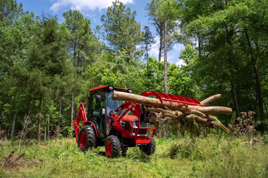 Red tractor with a front grapple attachment lifting several large tree logs in a green forest clearing under a blue sky with scattered clouds.