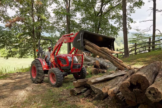 Man operating an orange tractor with front loader to move large logs in a wooded area with trees and a wooden fence.