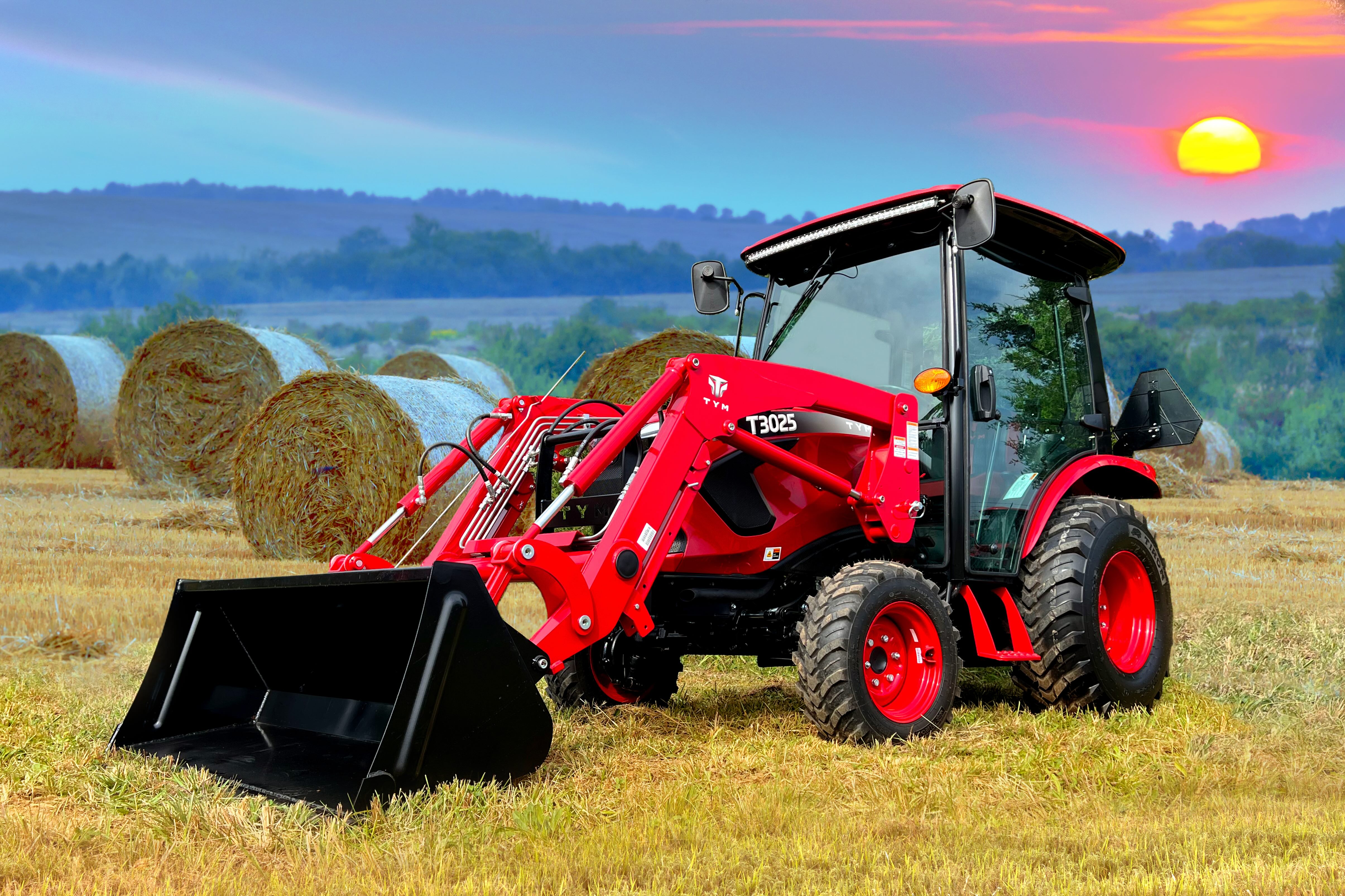 Red TYM T3025 tractor with front loader bucket parked on dry grass in a field with large round hay bales in the background under a colorful sunset sky.