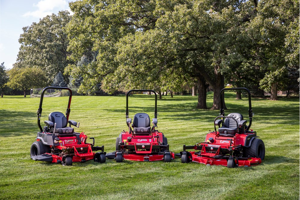 Three red zero-turn lawn mowers parked on green grass with large trees in the background under a partly cloudy sky.