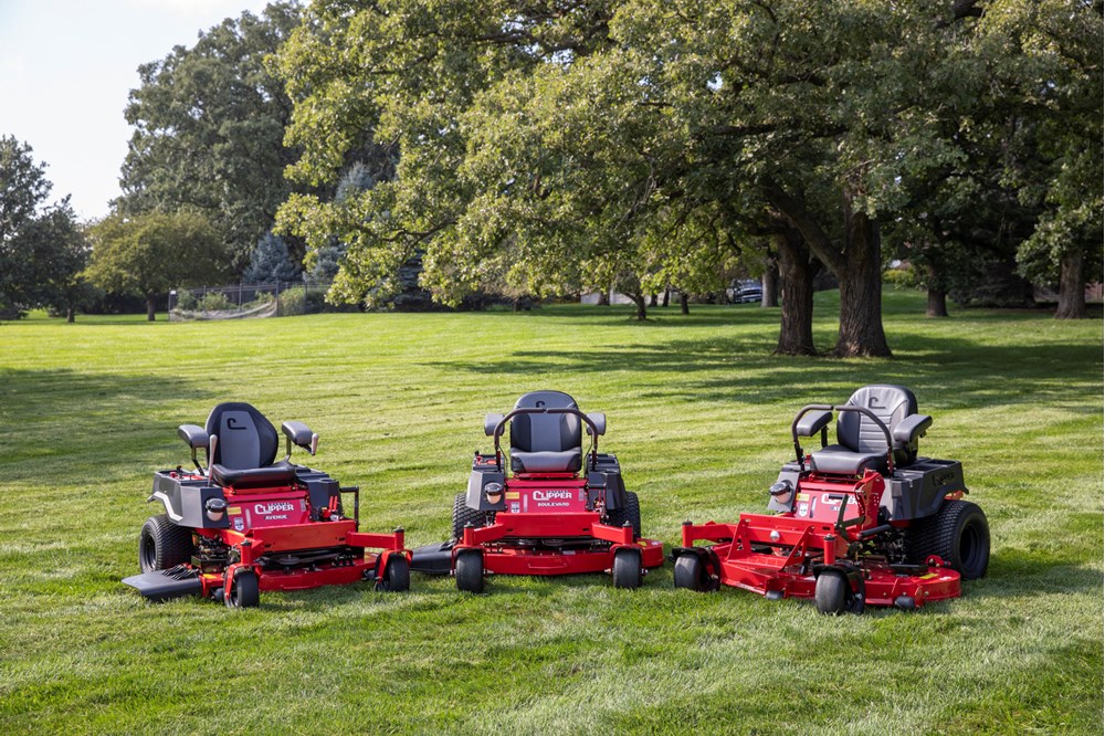 Three red Clipper zero-turn riding lawn mowers parked on green grass with large leafy trees in the background under a partly cloudy sky.