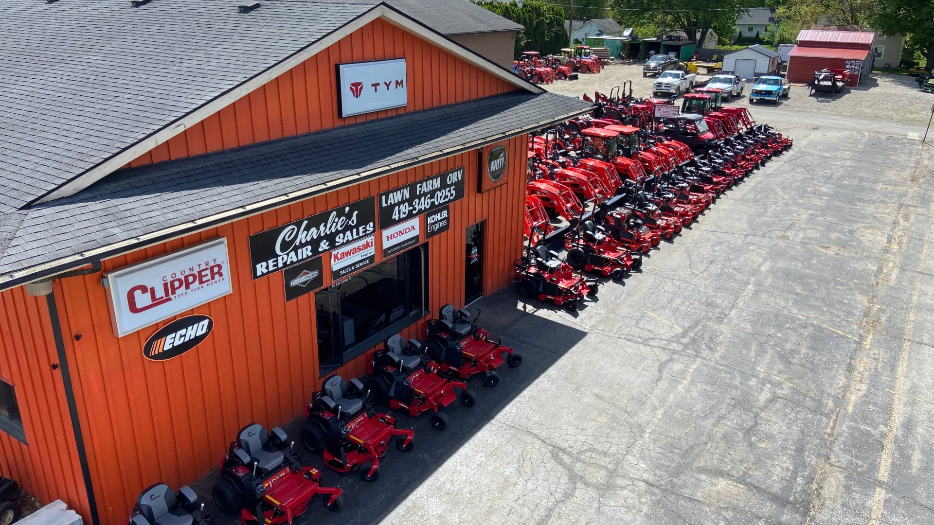 Large display of red lawn mowers and tractors lined up outside an orange building with multiple signs including 'Charlie's Repair & Sales,' 'Country Clipper,' Echo, Kawasaki, Honda, and Kohler Engines.