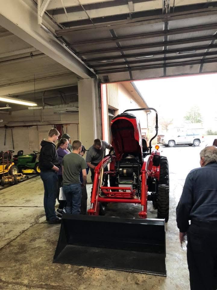 A group of people gathered around a red tractor with the engine hood open inside a garage or workshop, with some smaller green lawn tractors visible in the background and a white SUV outside.