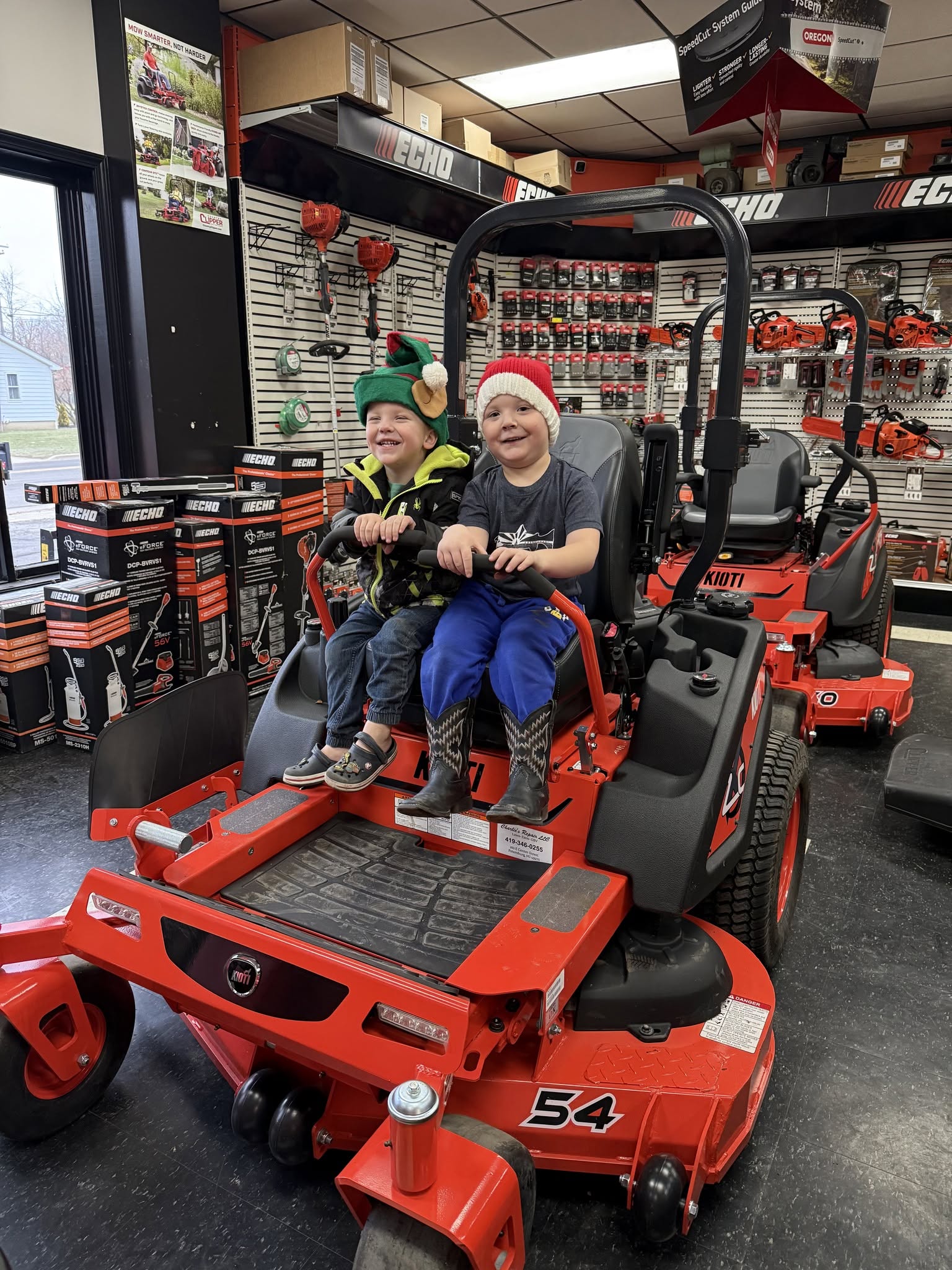 Two young boys sitting on a red Kioti riding lawn mower inside a store with lawn and garden equipment, both wearing festive hats and smiling.