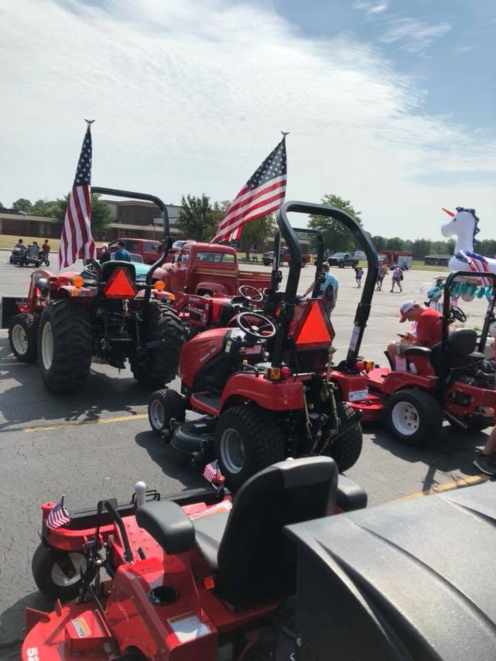 Red tractors and riding lawn mowers decorated with American flags parked on asphalt under blue sky, with people and vintage vehicles in the background at an outdoor event.