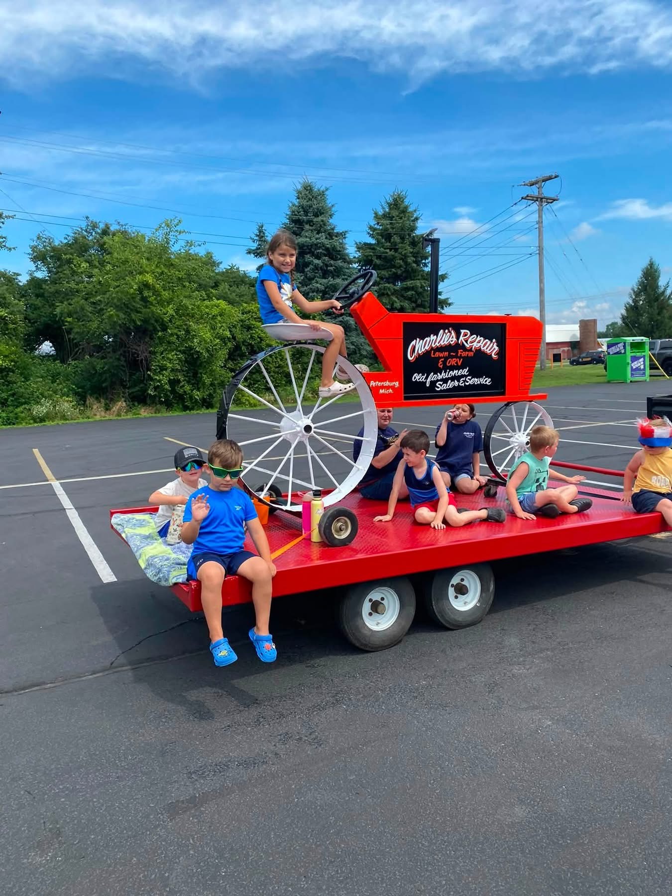 Seven children sitting and posing on a red flatbed trailer, with one girl sitting on a large red vintage-style tractor labeled 'Charlie's Repair Lawn - Farm & ORV Old fashioned Sales & Service' against a blue sky background.