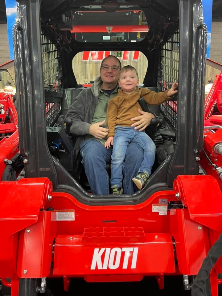 A man wearing glasses, a green shirt, and a gray jacket sits inside a red Kioti tractor holding a young boy dressed in a brown sweater and blue jeans, who is also sitting on his lap with his right arm resting on the tractor frame.