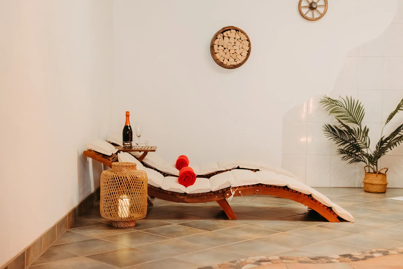 Entspannungsbereich mit zwei Holzbänken mit weißen Polstern und roten Handtüchern, einer Flasche Sekt mit Glas, einer Laterne mit brennender Kerze und einer Pflanze in einem Korb.