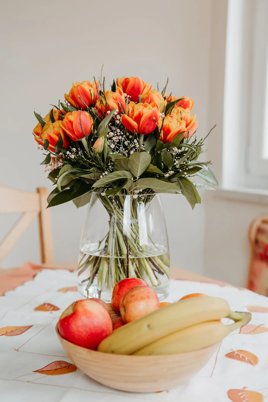 Vase mit orange-roten Tulpen und Schale mit Bananen und Äpfeln auf einem Tisch mit weißer Tischdecke.