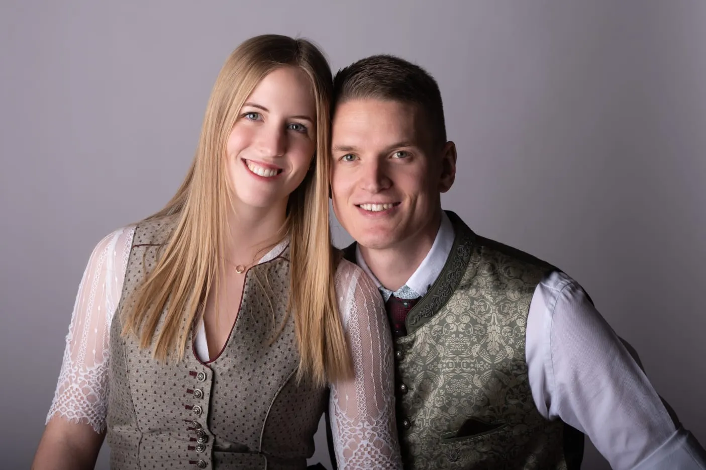 Paar in traditioneller Tracht lächelt vor grauem Hintergrund.