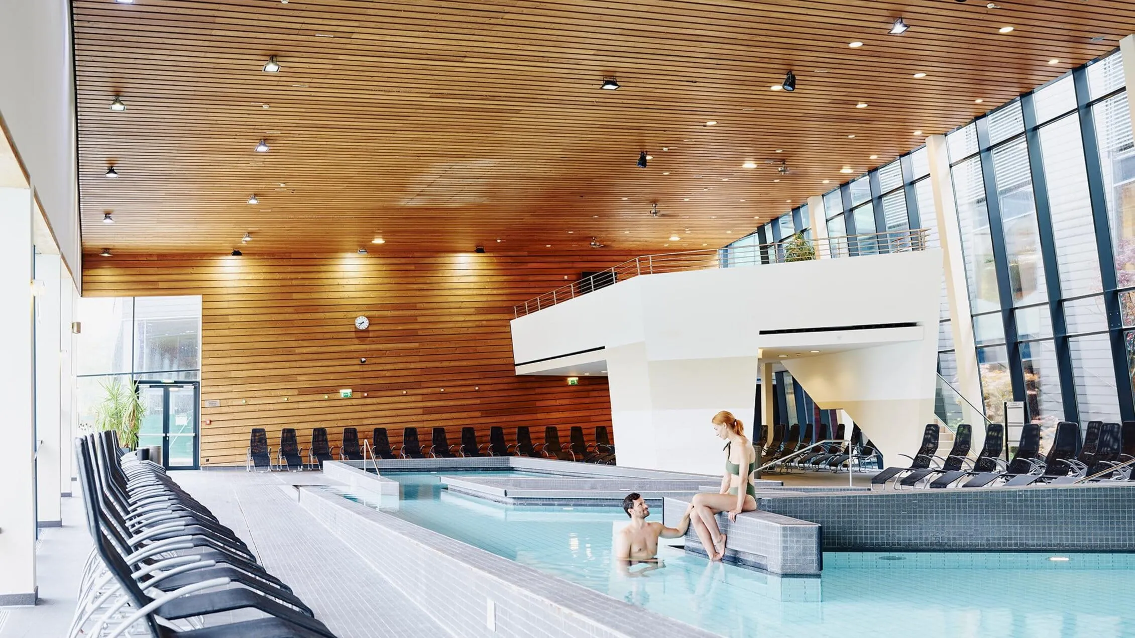 Innenansicht eines modernen Hallenbads mit Holzdecke, Glasfenstern und Liegestühlen, zwei Personen neben dem Becken.