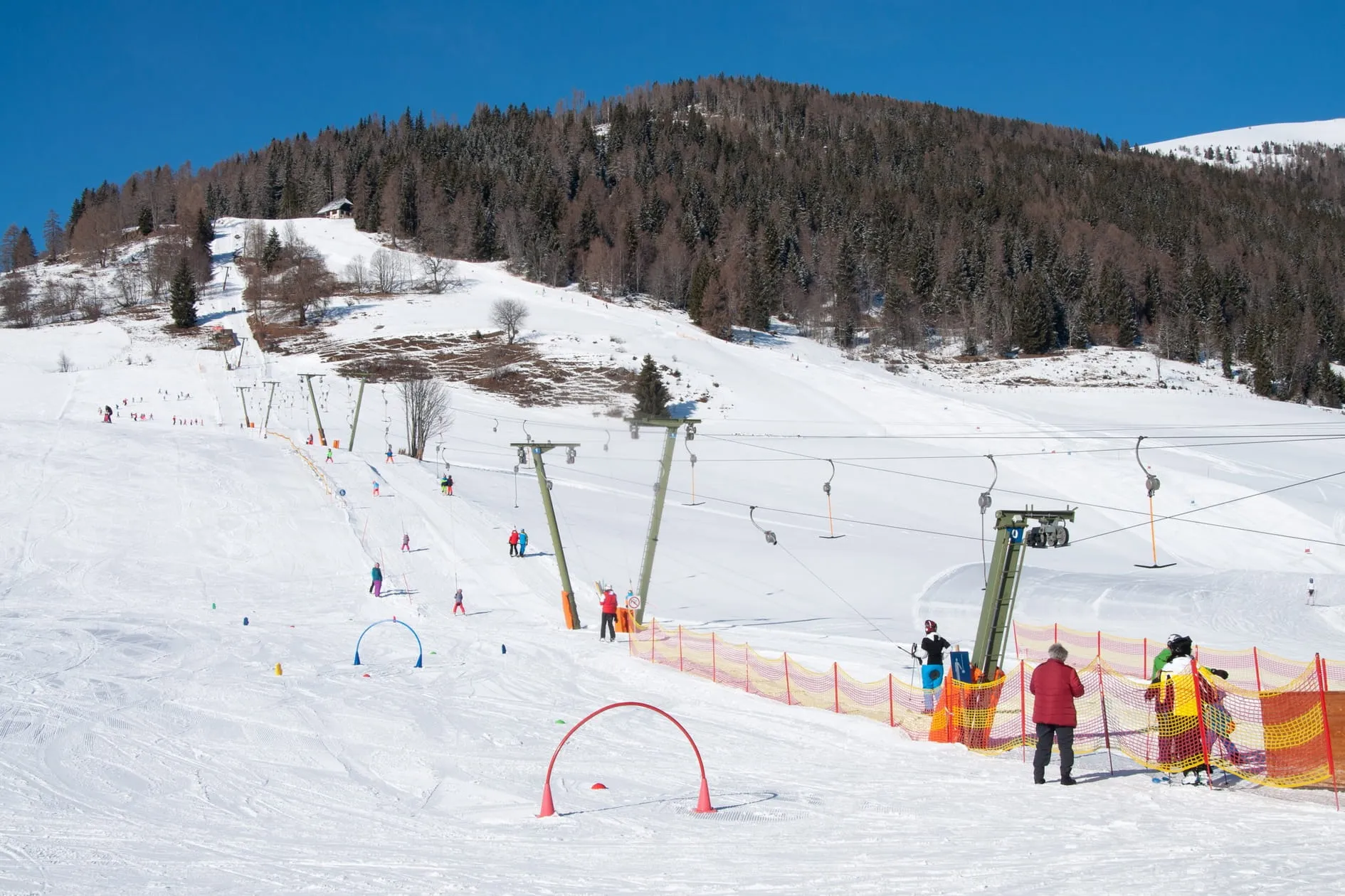 Skigebiet mit Skilift, mehreren Skifahrern auf schneebedeckter Piste und bewaldetem Hügel im Hintergrund unter blauem Himmel.