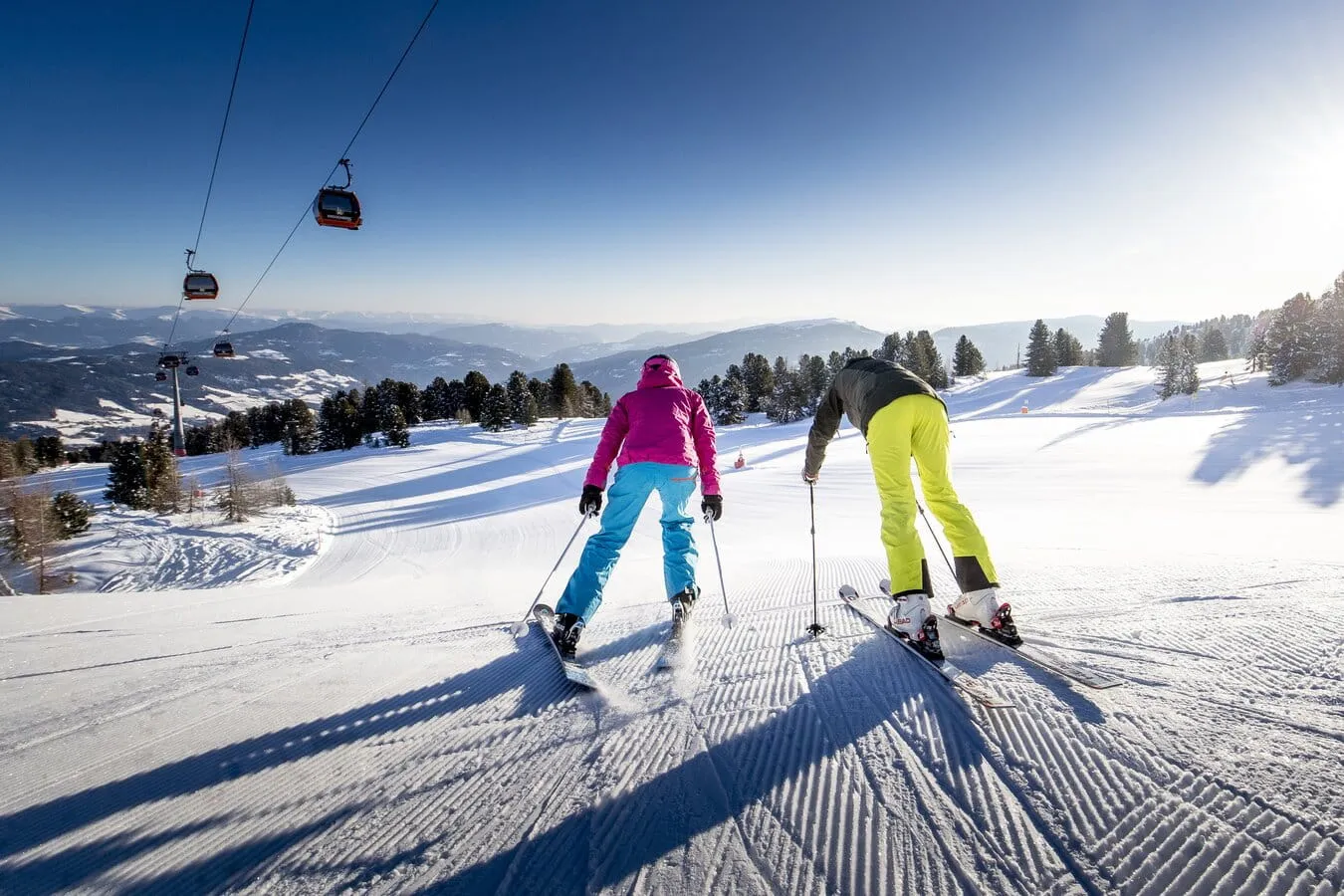 Zwei Skifahrer in bunter Kleidung fahren auf einer frisch präparierten Skipiste mit Blick auf verschneite Berge und Gondeln.