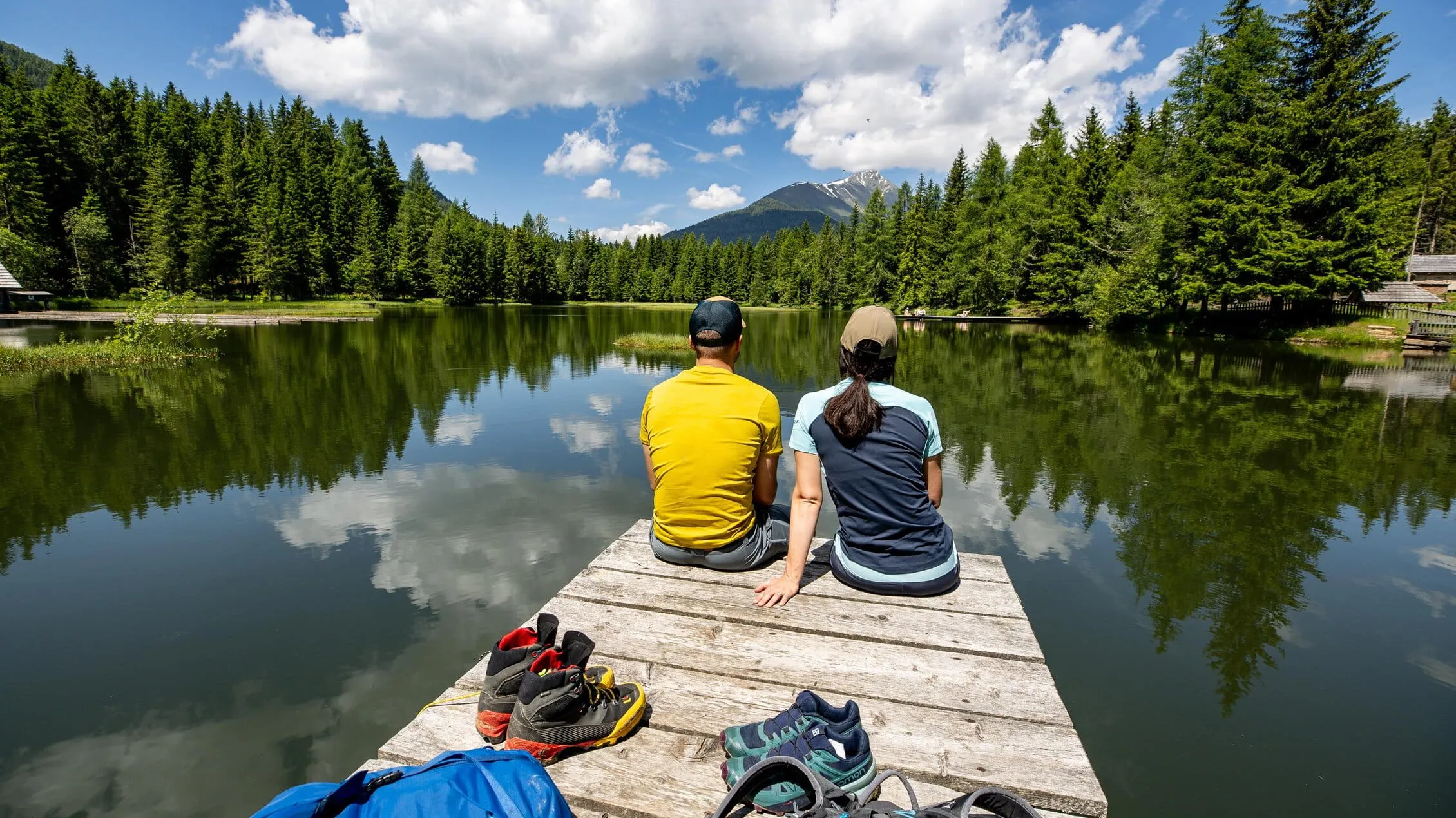 Ein Paar sitzt entspannt auf einem Holzsteg am See, umgeben von Wäldern und Bergen unter einem partly bewölkten Himmel.
