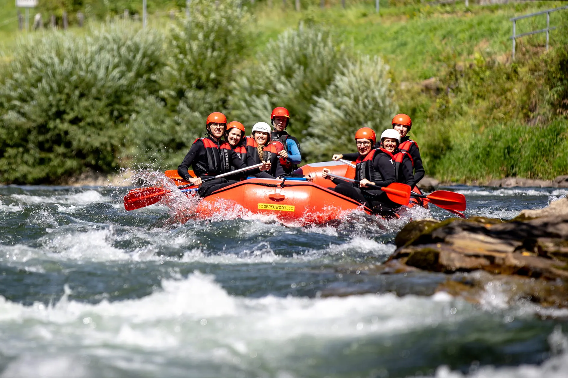 Gruppe von sieben Personen mit Helmen und Schwimmwesten beim Wildwasser-Rafting auf einem Fluss in einem roten Schlauchboot.