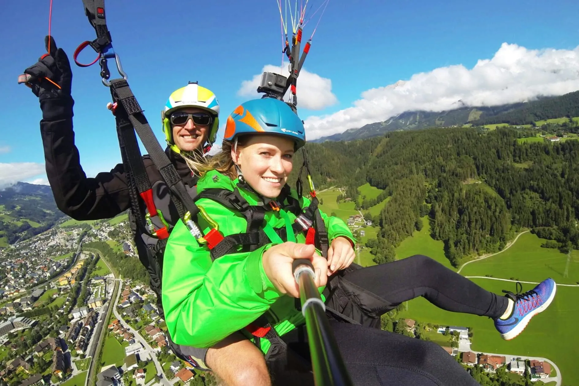 Zwei Personen beim Gleitschirmfliegen über einer grünen Landschaft mit Bergen und einem Dorf unter klarem Himmel.