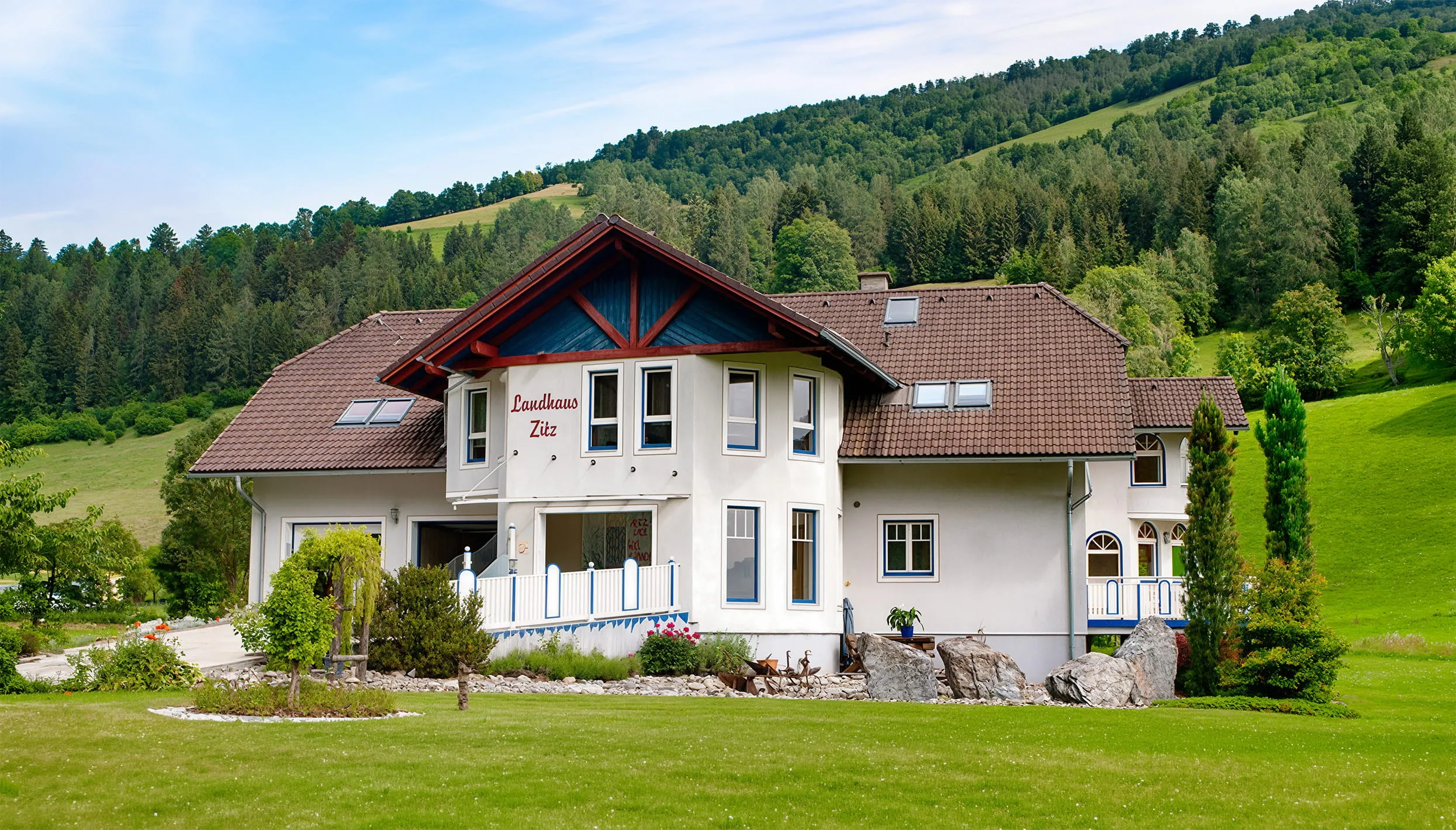 Weißes Landhaus mit braunem Dach vor bewaldeten Hügeln und grünem Rasen.